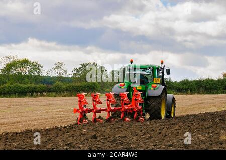 Green farm tractor working in field, pulling 5-furrow reversible plough,  stubble ploughing & turning soil (farmer driving) - North Yorkshire, GB. Stock Photo