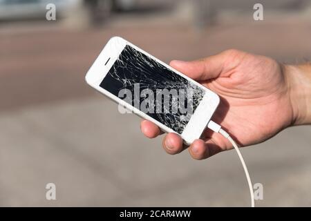 Male hand holding broken mobile smartphone after drop, closeup, outdoors, blurred background. Smartphone with broken (cracked) screen. Picking broken Stock Photo