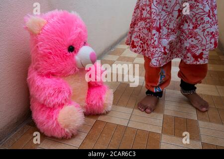 a close shot of pink teddy bear sitting on floor with baby girl legs background Stock Photo