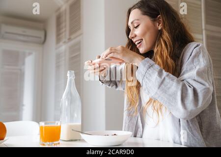 Image of cheerful beautiful woman taking photo on smartphone while having breakfast in white kitchen Stock Photo