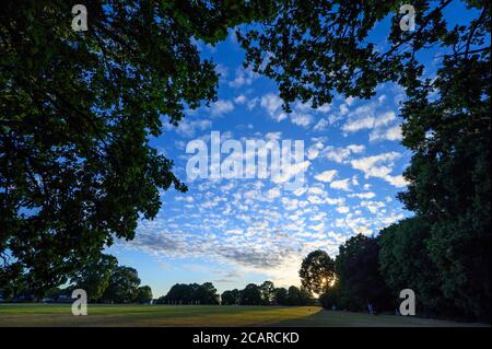 Sunset in Harvington Park, Beckenham, Kent. Small fluffy altocumulus clouds against a blue sky with the setting sun. Unidentifiable people in the park Stock Photo