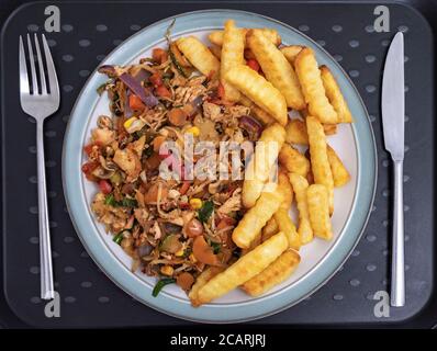 Closeup POV overhead shot of a knife and fork next to a dinner plate on a tray, of hot stir fry salmon and vegetables, accompanied by chips / fries. Stock Photo