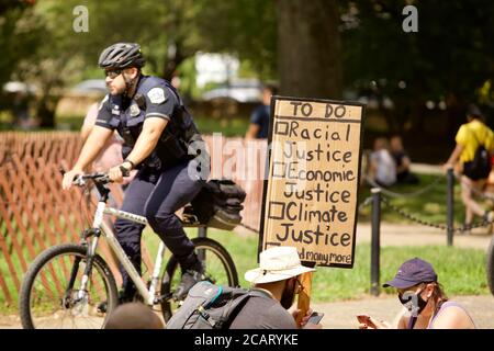 Washington, DC, USA. 8th Aug, 2020. Pictured: Protester holds a sign listing the social and legal changes needed as one of the 10  Metropolitan Police (DC police) rides by on a bike. Credit: Allison C Bailey/Alamy. Credit: Allison Bailey/Alamy Live News Stock Photo