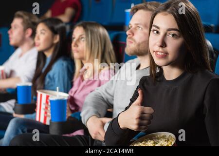 Best friends time. Beautiful brunette showing thumbs up while enjoying movies with her friends at the local movie theatre Stock Photo