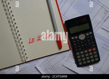 Closeup message kill the bills written with red pen on spiral notebook next to calculator against the bills in background Stock Photo