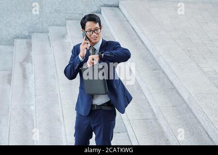 Handsome positive young Asian entrepreneur checking time on his wristwatch when talking on phone with colleague Stock Photo