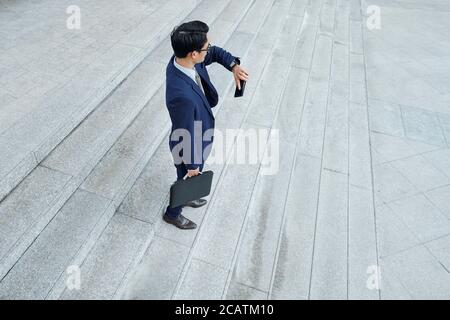 Young businessman waiting for colleague outdoors and checking time on smartwatch Stock Photo