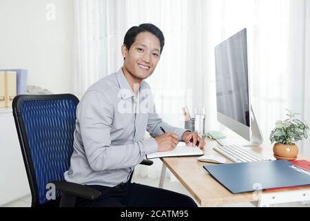 Portrait of handsome young Vietnamese entrepreneur working from home at his desk and taking notes in planner Stock Photo