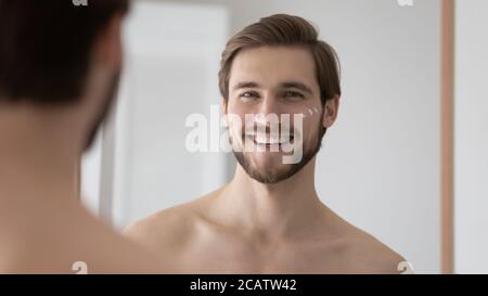 Head shot mirror reflection smiling young man applying moisturizing cream Stock Photo