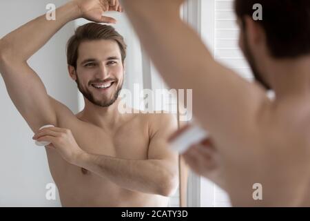 Mirror reflection smiling young man applying antiperspirant on armpit Stock Photo