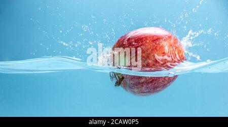 Ripe red apple is falling into clear water, forming lots of splashes. Concept of tasty juicy fruits rich in vitamins. Stock Photo