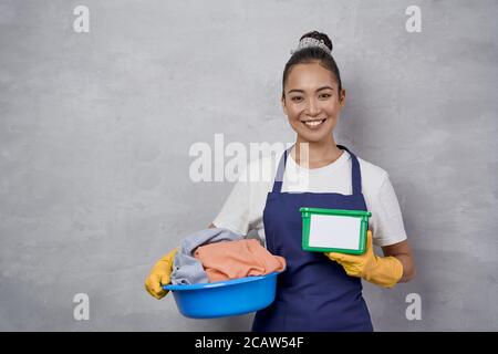 Doing laundry. Portrait of happy maid woman in uniform holding basket with laundry and green plastic box with washing capsules, smiling at camera against grey wall. Housekeeping, housework Stock Photo