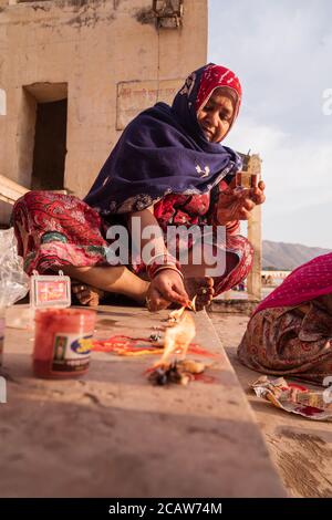 Pushkar / India - March 10, 2020: Indian women from Rajasthan wearing red traditional clothes sitting in stairs near Pushkar sacred lake making offering with candle Stock Photo