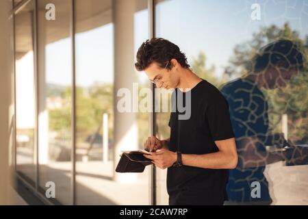 Young man using digital tablet while standing against glass window at college campus Stock Photo