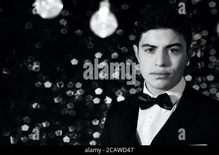 Handsome latino young man wearing tuxedo standing under string of lights with bokeh blurred lights in the background and room for copy Stock Photo