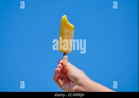 yellow popsicle in the hand of a against blue sky with copy space Stock Photo