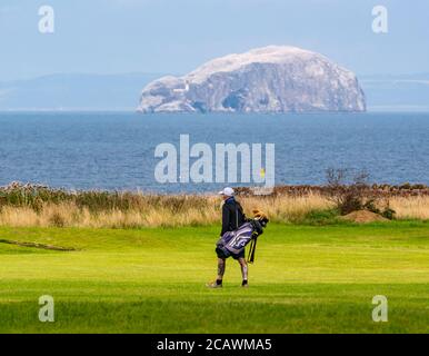 East Lothian, Scotland, United Kingdom, 9th August 2020. UK Weather:  sunny Summer day for outdoor sports. A golfer with tattoos carrying a Callaway golf bag with clubs on Winterfield golf course in Dunbar with the Bass Rock gannet colony on the horizon Stock Photo