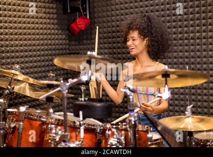 Laughing happy vivacious young Afro American woman in her twenties seated playing drums and cymbals in a studio Stock Photo