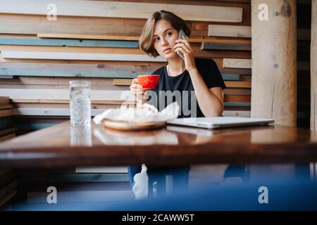Busy young woman drinking coffee in a cafe while talking on the phone