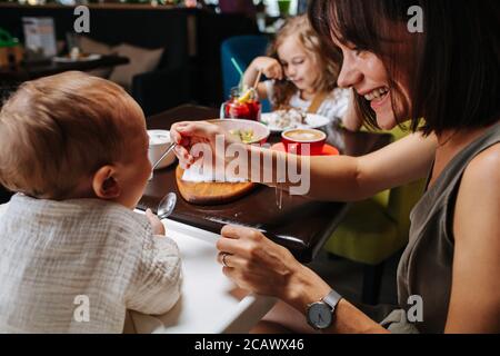 Mom feeding her baby, her daughter eats on the other side of the table. Stock Photo
