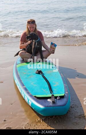 Young woman sitting on paddleboard, paddle board, with cockapoo dog sitting on her lap at Poole, Dorset, UK on hot sunny day in August Stock Photo