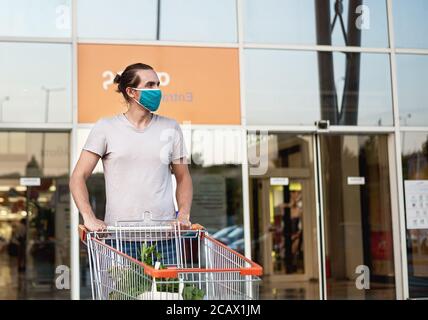 Young man wearing protective medical mask and carrying a shopping cart with grocery inside Stock Photo