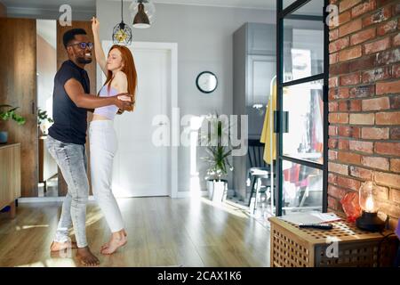 romantic funny couple dancing at home, attractive redheadyoung woman and handsome african man are enjoying spending time together Stock Photo