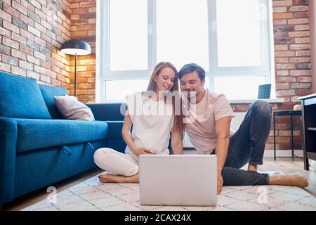 sweet pregnant woman and man surf the net sitting on the floor with laptop, at home in domestic wear Stock Photo