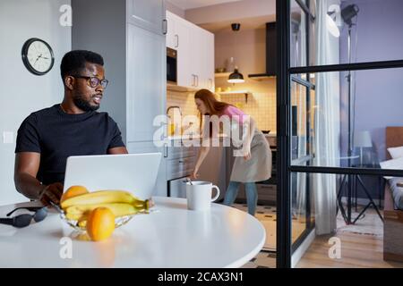 interracial couple in the kitchen at home, african guy sit using laptop, work from home, redhead female doing household chores in the background Stock Photo