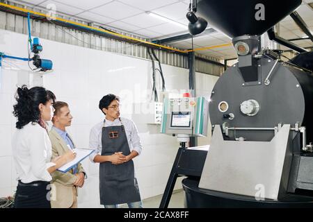 Small coffee roastery owner in apron telling about new modern facilities like roaster to investors or fire inspectors Stock Photo