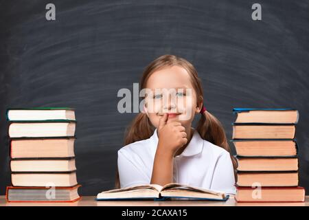 Schoolgirl little girl sitting at a table on the background of a school black chalkboard. Child thoughtful among the stacks of books. Education concep Stock Photo