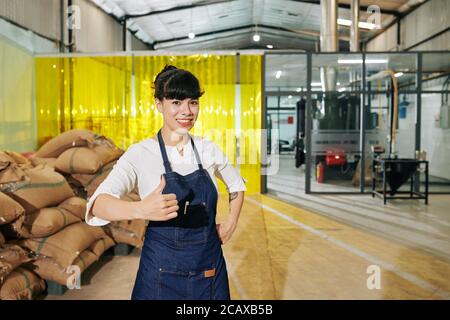 Portrait of happy excited female coffee roastery owner showing thumbs-up and looking at camera Stock Photo