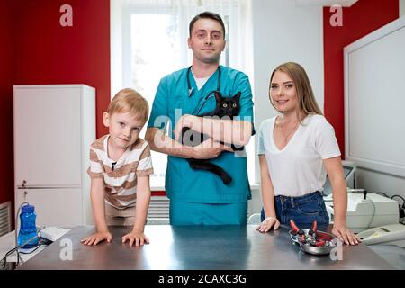 portrait of vet and cat owners in hospital, careful affable veterinarian hold black cat in hands and look at camera, woman and child boy stand near, s Stock Photo