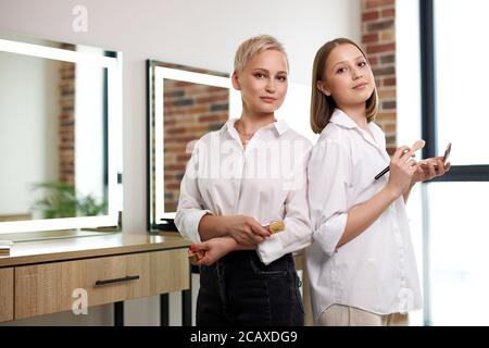 portrait of confident caucasian make-up artists in beauty studio, ladies in formal wear hold cosmetics in hands, stand back to back, colleagues Stock Photo