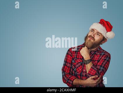 Bearded handsome hipster young man in Christmas hat looking up posing in studio thinking of gift ideas isolated on blue studio wall background with co Stock Photo