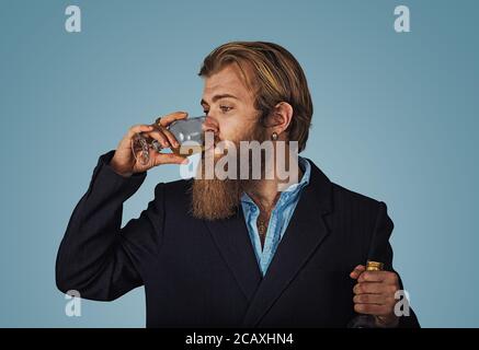 Handsome bearded well-dressed man in jacket holding tasting drinking glass of beverage whiskey alcohol isolated on grey gray studio background wall. d Stock Photo