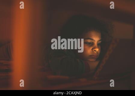 Little girl aged 8 years old is using her digital Tablet in her bed. She is being illuminated by the brightness of the screen only. Stock Photo