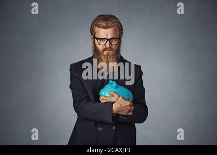 Young formal man in glasses extremely fascinated with money saving holding with greedy hand gesture the piggy bank. Bearded hipster businessman. Isola Stock Photo
