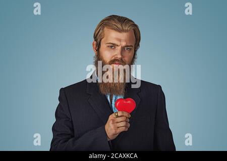 Serious Bearded hipster businessman man in black jacket and blue shirt holding red heart standing holding red heart shaped gift box and looking at cam Stock Photo
