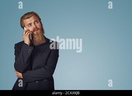 Bearded pensive hipster young businessman holding looking up talking speaking on phone thinking isolated on blue studio wall background. Serious face Stock Photo
