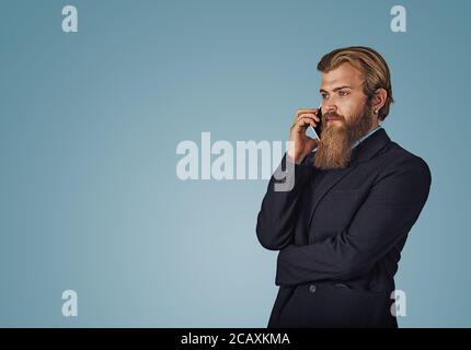 Bearded hipster business man holding talking by mobile phone isolated on blue studio wall Background. Serious face expression, human emotion, body lan Stock Photo