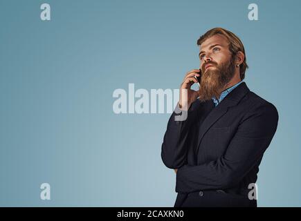 Bearded pensive hipster young businessman male holding cellphone looking up talking speak on phone thinking Isolated on blue Background. Serious face Stock Photo