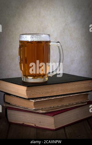 A glass of golden beer over a stack of hardbound books on a wooden table against grey background, side view Stock Photo