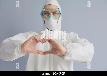 Doctor in hazmat suit showing heart with hands on grey background. Medic encouraging donation to help fight epidemic Stock Photo
