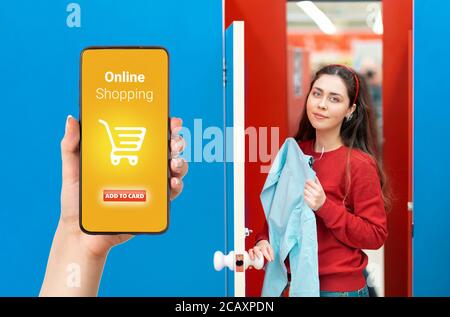 A person's hand holds a mobile phone, with a shopping basket on the screen. In the background, a woman stands with clothes in a fitting room. The conc Stock Photo