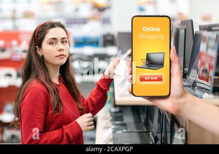 A person's hand holds a mobile phone, with a shopping basket on the screen. In the background, a pretty woman selects digital products. The concept of Stock Photo