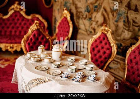 Close-up of three types of tea set on a table with lace tablecloth and chairs with gold rims and red trim. Stock Photo