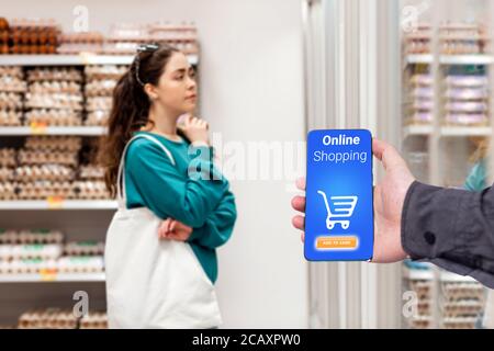 A person's hand holds a mobile phone, with a shopping basket on the screen. In the background, a woman selects products, in a blur. The concept of onl Stock Photo