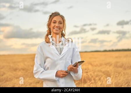 Woman farmer white coat smart farming standing farmland smiling using digital tablet Female agronomist specialist research monitoring analysis data ag Stock Photo