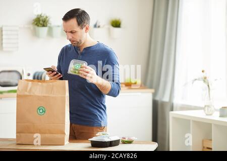 Waist up portrait of mature bearded man using smartphone while unpacking food delivery bag in kitchen interior, copy space Stock Photo